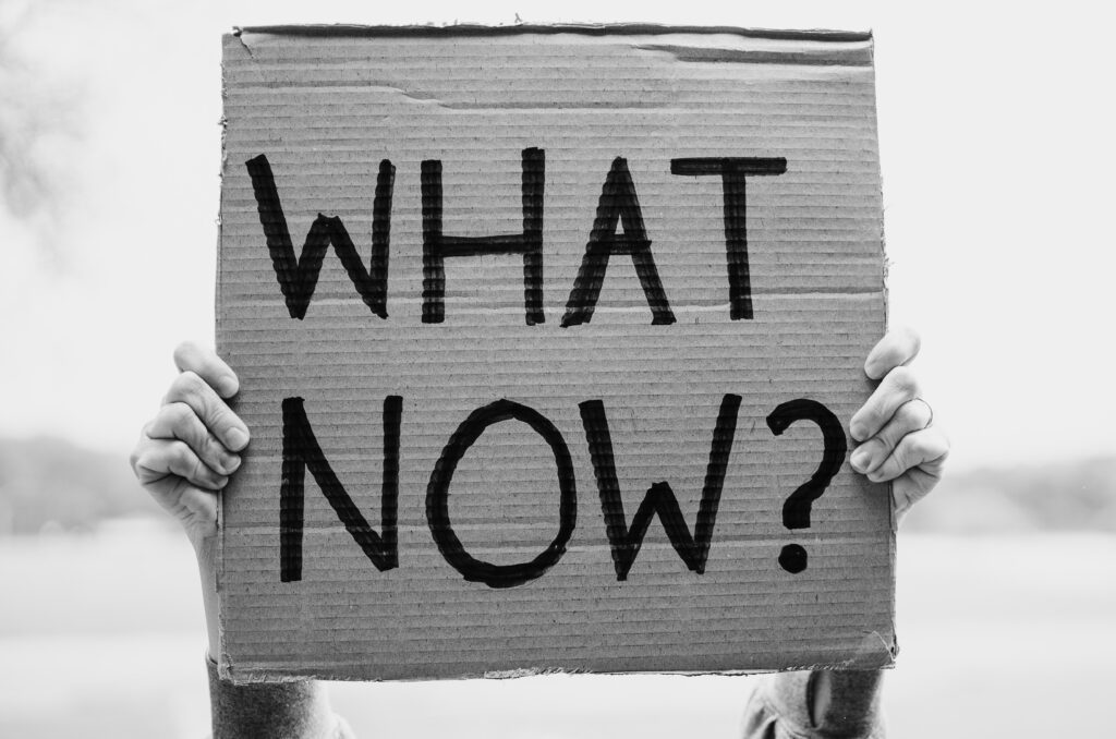 A black and white image showing two hands holding up a cardboard banner which says 'what now?'