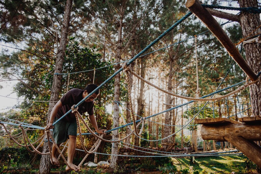 A man with bare feet is making his way across a rope bridge in a forest with trees surrounding him and golden sunlight peeping through. Deceptively relaxing in my opinion.