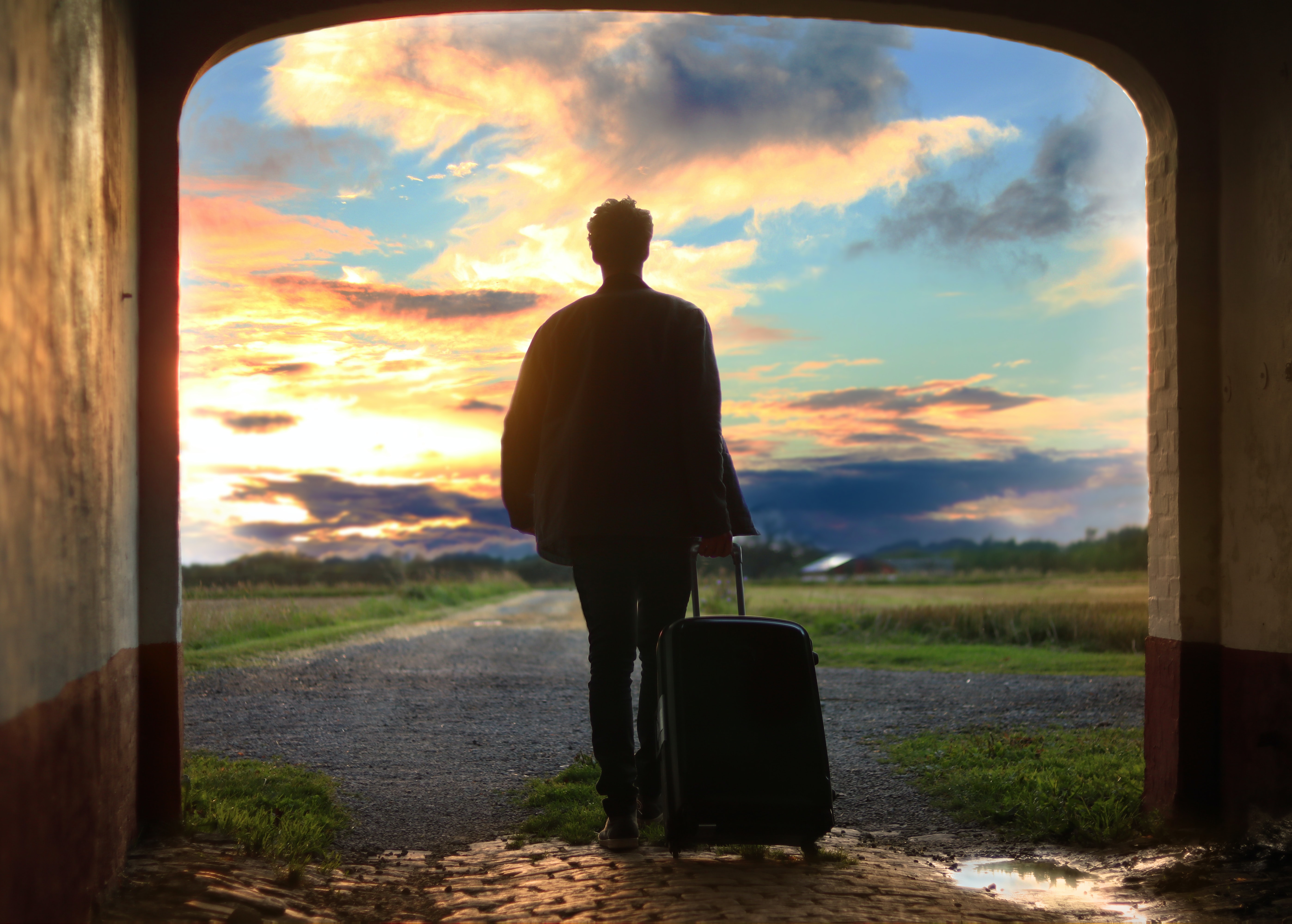 man with suitcase walking toward a path with a sunset in front of him