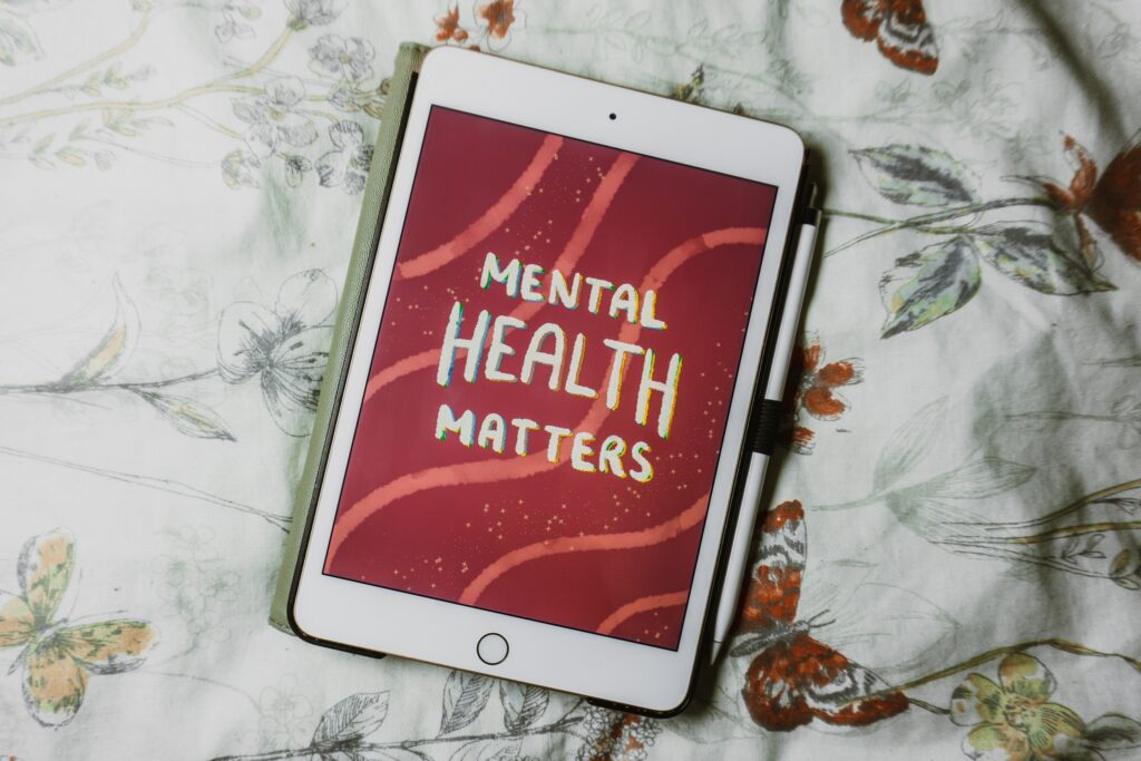 an ipad screen shows a graphic that says 'mental health matters' in white text with a red background and pink wavy lines. The ipad is on a white duvet cover that has yellow and red butterflies