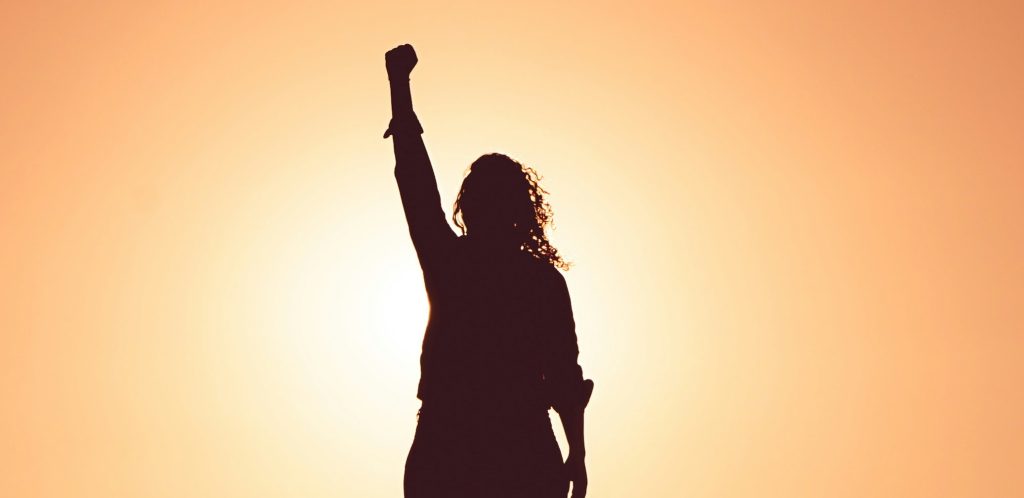 silhouette of woman with long hair raising her fist to the air. Orange sky in background