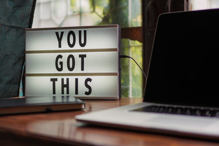 A laptop and a book on a desk with a neon sign that says you got this