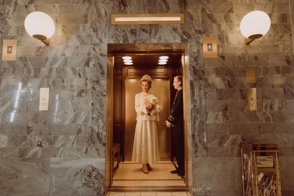 An elegant white woman in a white dress with a white dog stands in a gold and wood elevator next to a man in uniform. The wall of the corridor appears to be grey marble. 