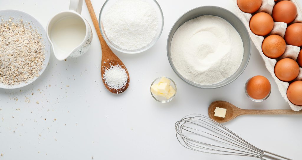 A white surface with spoons, a whisk, a jug of milk, bowls with butter, oats, and flour, and a carton of eggs