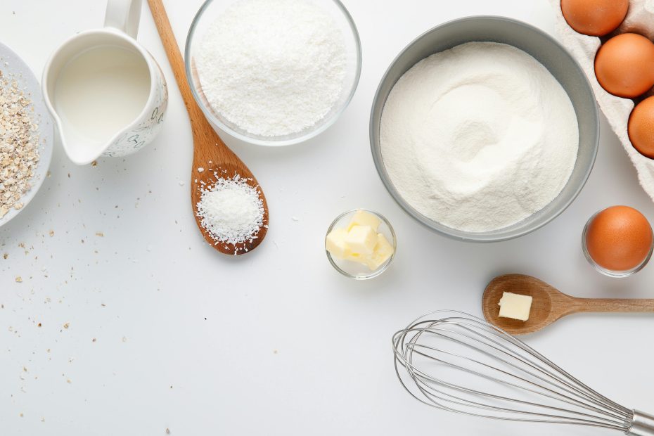 A white surface with spoons, a whisk, a jug of milk, bowls with butter, oats, and flour, and a carton of eggs